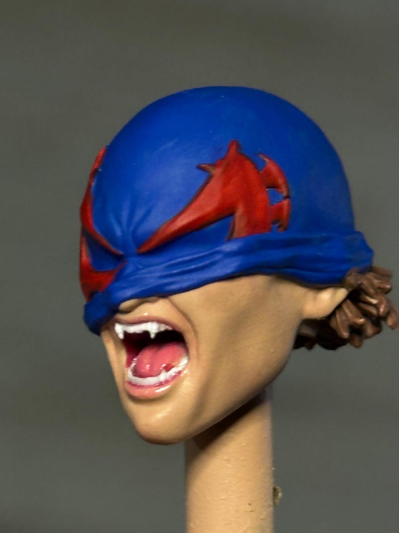 May include: A sculpted head model featuring a blue mask with red wing-like accents covering the eyes. The mouth is open in a wide expression, revealing white fangs and a pink tongue. The skin tone is light tan, and brown hair is visible at the back.