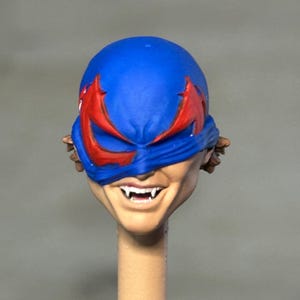 May include: A sculpted action figure head featuring a blue mask with red eye accents, revealing a tan-skinned face with a wide, toothy grin and visible fangs. The head is mounted on a thin, cylindrical beige post, showing brown hair at the sides.