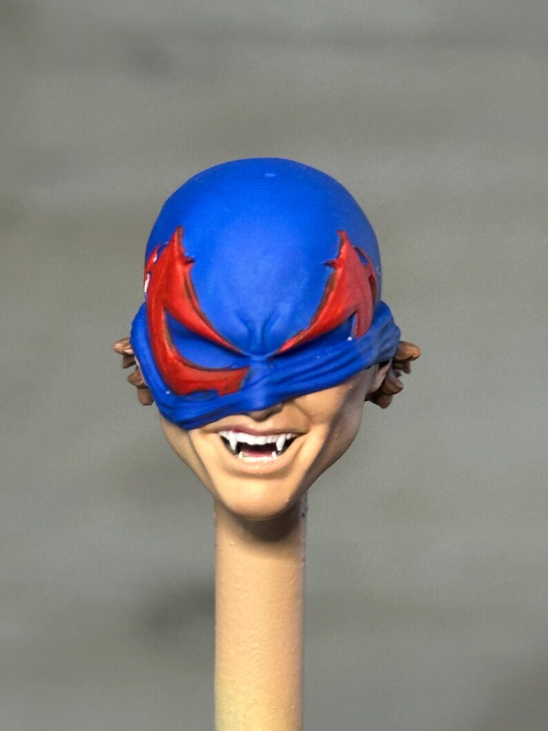 May include: A sculpted action figure head featuring a blue mask with red eye accents, revealing a tan-skinned face with a wide, toothy grin and visible fangs. The head is mounted on a thin, cylindrical beige post, showing brown hair at the sides.