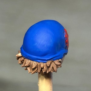 May include: A miniature figurine head featuring a smooth, vibrant blue cap worn over short, textured light brown hair. A small, circular red emblem with a detailed spiderweb pattern is visible on the side of the blue cap, adding a distinct accent.