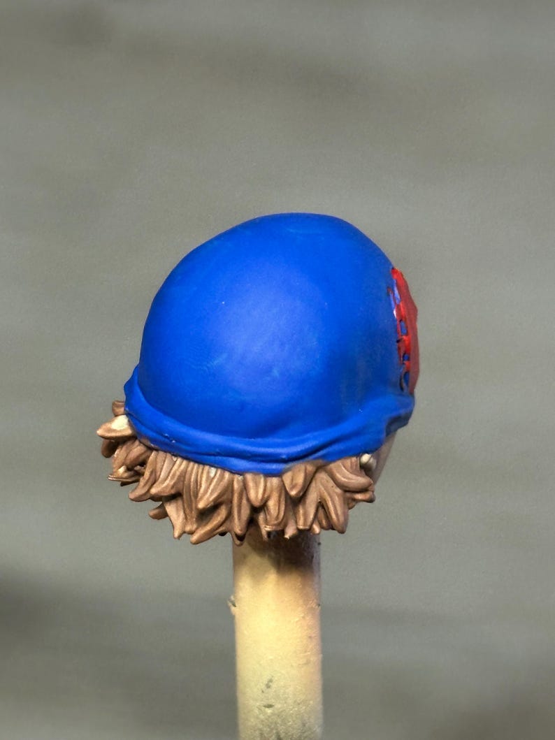 May include: A miniature figurine head featuring a smooth, vibrant blue cap worn over short, textured light brown hair. A small, circular red emblem with a detailed spiderweb pattern is visible on the side of the blue cap, adding a distinct accent.