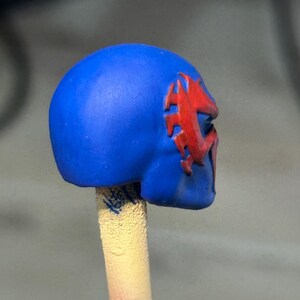 May include: A blue helmet-like object featuring a raised red spider-shaped emblem on the side. The object is mounted on a cylindrical beige stick, showing a smooth matte finish on the blue surface and a textured appearance on the support stick.