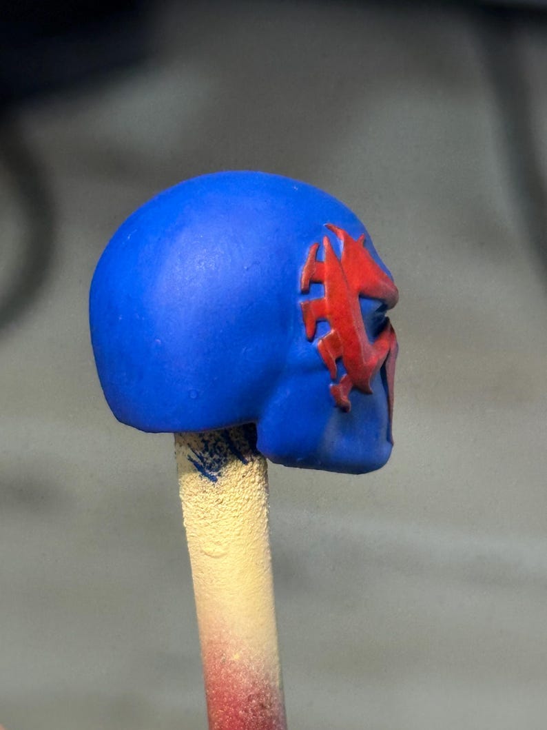May include: A blue helmet-like object featuring a raised red spider-shaped emblem on the side. The object is mounted on a cylindrical beige stick, showing a smooth matte finish on the blue surface and a textured appearance on the support stick.