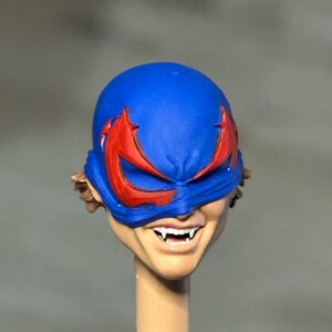 May include: A miniature action figure head featuring a bright blue mask with sharp red eye markings. The face has a wide, toothy grin revealing pointed white fangs. Brown hair is visible at the sides, and the head is mounted on a thin, cylindrical post.