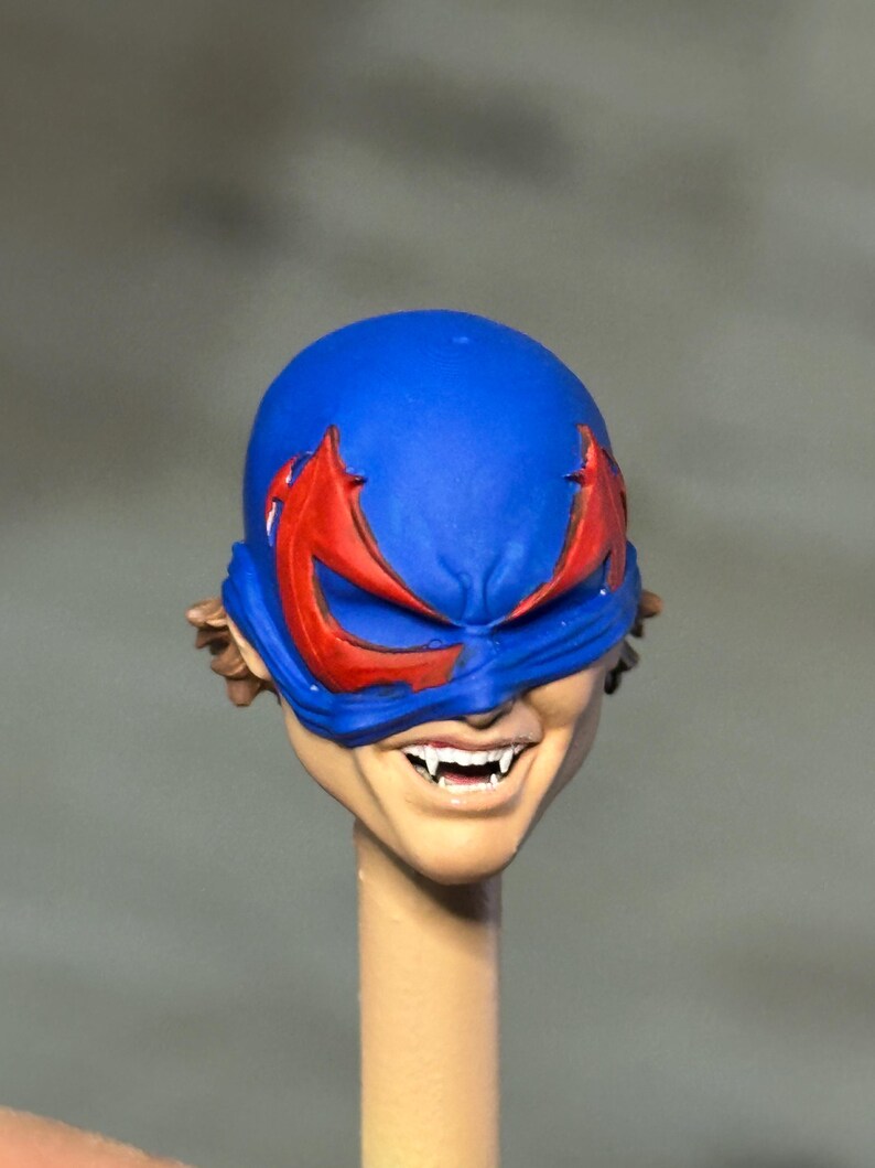 May include: A miniature action figure head featuring a bright blue mask with sharp red eye markings. The face has a wide, toothy grin revealing pointed white fangs. Brown hair is visible at the sides, and the head is mounted on a thin, cylindrical post.
