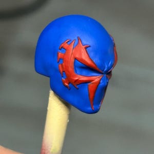 May include: A small, sculpted head piece featuring a vibrant blue base color with a prominent, raised red spider-like emblem extending across the eye area. The object is mounted on a thin, cylindrical stick, showcasing detailed craftsmanship.