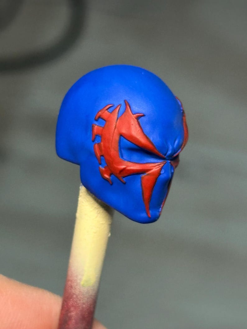 May include: A small, sculpted head piece featuring a vibrant blue base color with a prominent, raised red spider-like emblem extending across the eye area. The object is mounted on a thin, cylindrical stick, showcasing detailed craftsmanship.