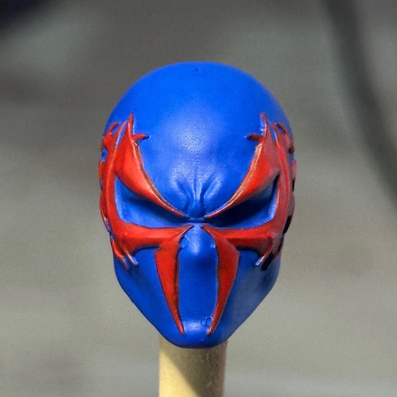 May include: A blue sculpted head piece featuring a prominent red spider-like emblem that extends from the eye sockets down to the nose area. The surface has a matte finish with molded details around the eyes and forehead, creating a distinct mask.