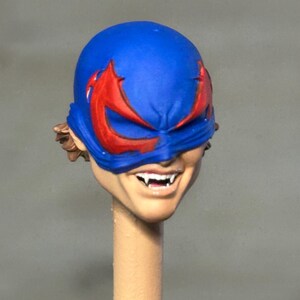 May include: A miniature sculpted head featuring a bright blue mask with red eye accents. The face has a visible grin showing sharp white fangs, and brown hair is visible at the sides. The head is mounted on a thin, light-colored cylindrical post.