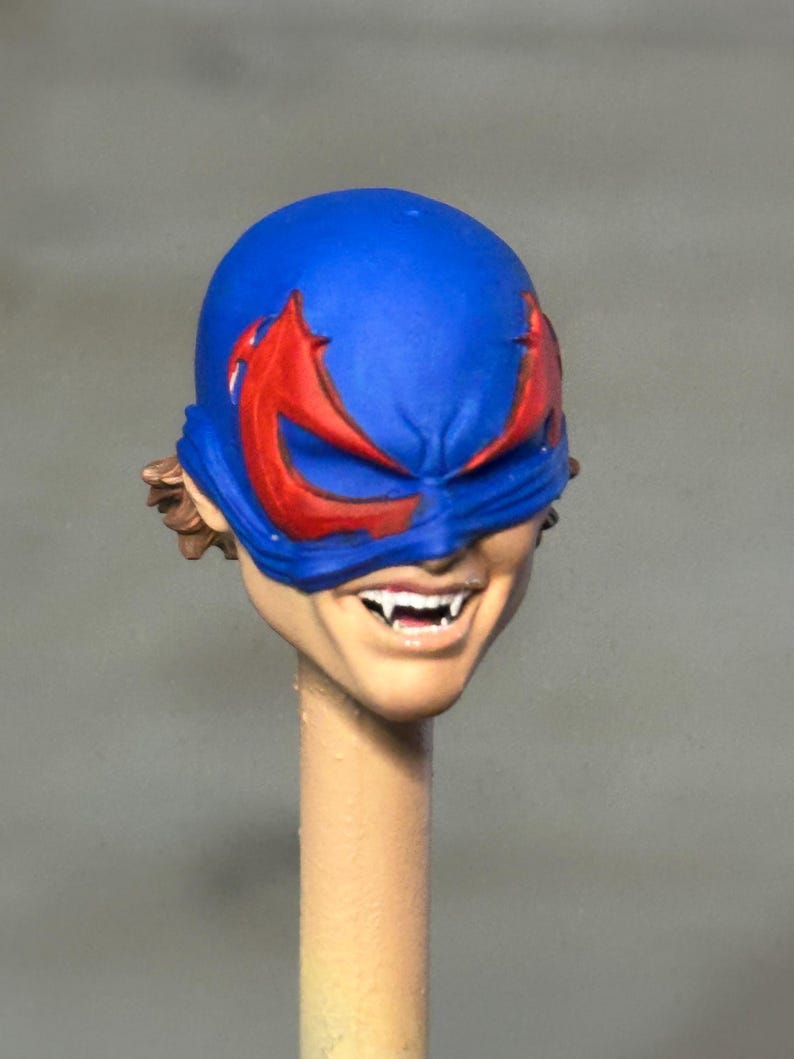 May include: A miniature sculpted head featuring a bright blue mask with red eye accents. The face has a visible grin showing sharp white fangs, and brown hair is visible at the sides. The head is mounted on a thin, light-colored cylindrical post.