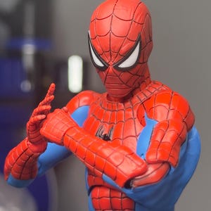 May include: A red and blue Spider-Man action figure with a detailed web pattern on the costume. The figure is in a dynamic pose, with arms crossed and hands gesturing. The eyes are white, and the figure has a black spider emblem on the chest.