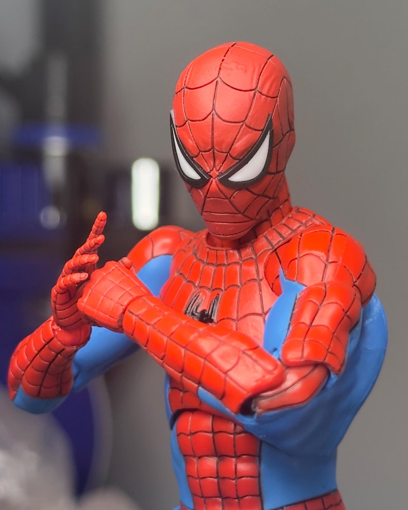 May include: A red and blue Spider-Man action figure with a detailed web pattern on the costume. The figure is in a dynamic pose, with arms crossed and hands gesturing. The eyes are white, and the figure has a black spider emblem on the chest.