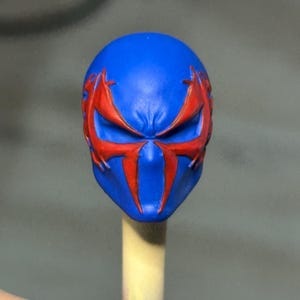 May include: A blue sculpted head piece featuring a prominent red spider-like emblem that extends across the eye sockets and down the center of the face. The design includes sharp, angular lines and a smooth, matte finish on the blue surface.