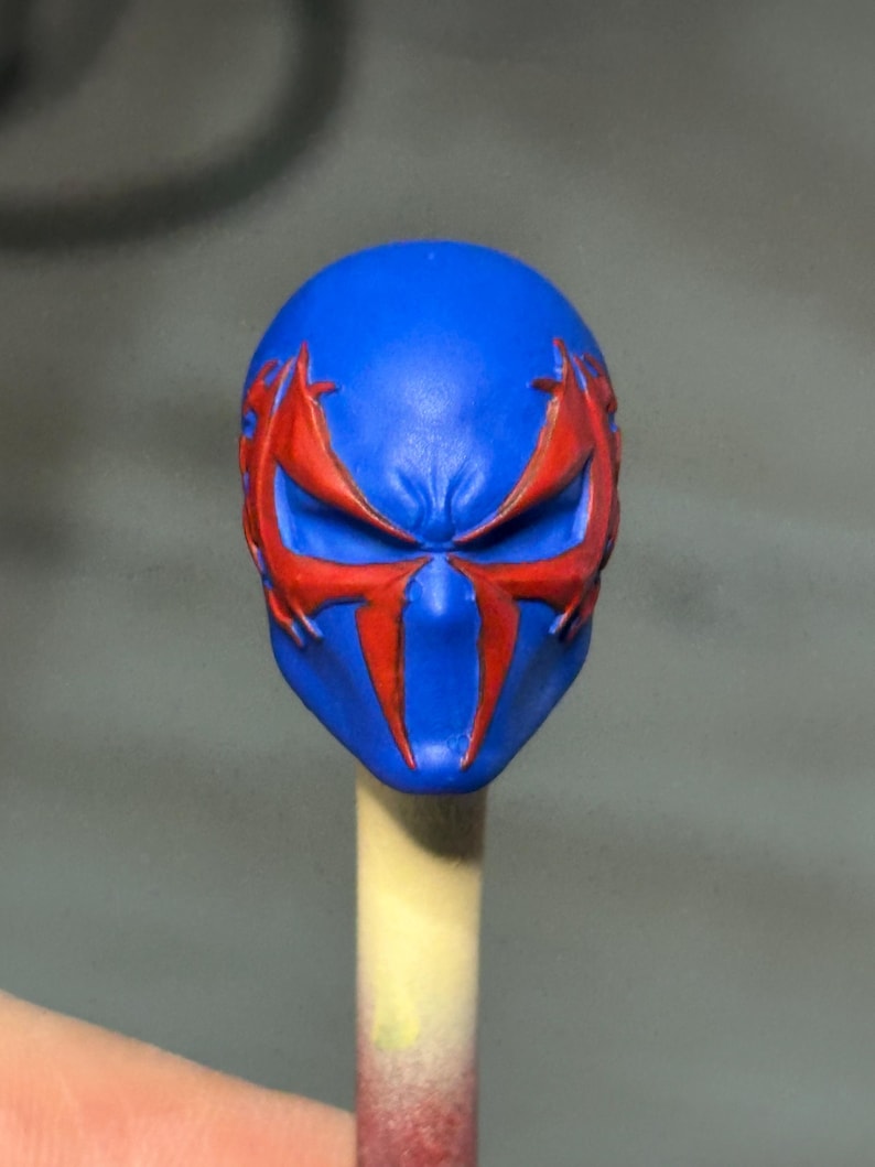 May include: A blue sculpted head piece featuring a prominent red spider-like emblem that extends across the eye sockets and down the center of the face. The design includes sharp, angular lines and a smooth, matte finish on the blue surface.