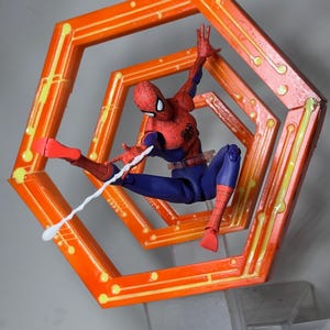 May include: A Spider-Man action figure in a dynamic pose, wearing a red and blue suit, shooting a white web. The figure is set against an orange, geometric frame with yellow accents, mounted on a clear stand.