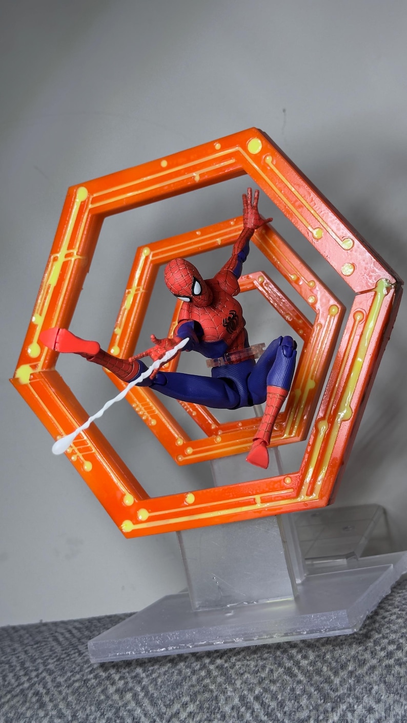 May include: A Spider-Man action figure in a dynamic pose, wearing a red and blue suit, shooting a white web. The figure is set against an orange, geometric frame with yellow accents, mounted on a clear stand.