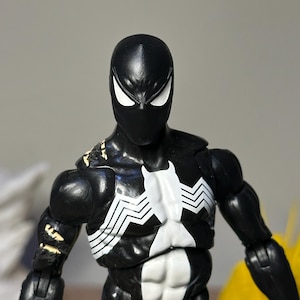 May include: A black action figure featuring a white spider emblem on the chest and white eye lenses. The figure has a muscular build with visible articulation points at the shoulders and arms, and shows signs of wear with small areas of paint loss.