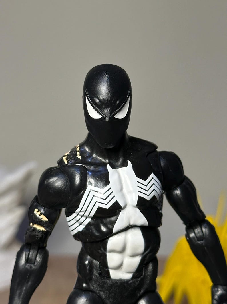 May include: A black action figure featuring a white spider emblem on the chest and white eye lenses. The figure has a muscular build with visible articulation points at the shoulders and arms, and shows signs of wear with small areas of paint loss.