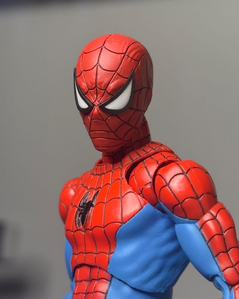May include: A red and blue Spider-Man action figure. The figure has a red mask with white eye lenses and a black spider emblem on the chest. The figure's costume is red with blue accents.