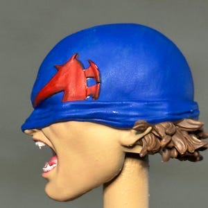 May include: A sculpted figurine head featuring a bright blue helmet with a prominent red emblem on the side. The face is depicted in profile with an open mouth showing teeth, and brown hair is visible at the base of the helmet. The skin is a light tan.