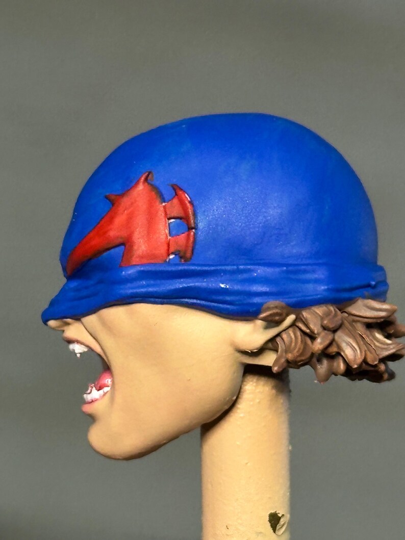 May include: A sculpted figurine head featuring a bright blue helmet with a prominent red emblem on the side. The face is depicted in profile with an open mouth showing teeth, and brown hair is visible at the base of the helmet. The skin is a light tan.