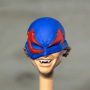 May include: A sculpted action figure head featuring a bright blue mask with red eye accents. The face has a light skin tone, a wide grin, and visible white fangs protruding from the mouth. Brown hair is partially visible on the sides of the head.