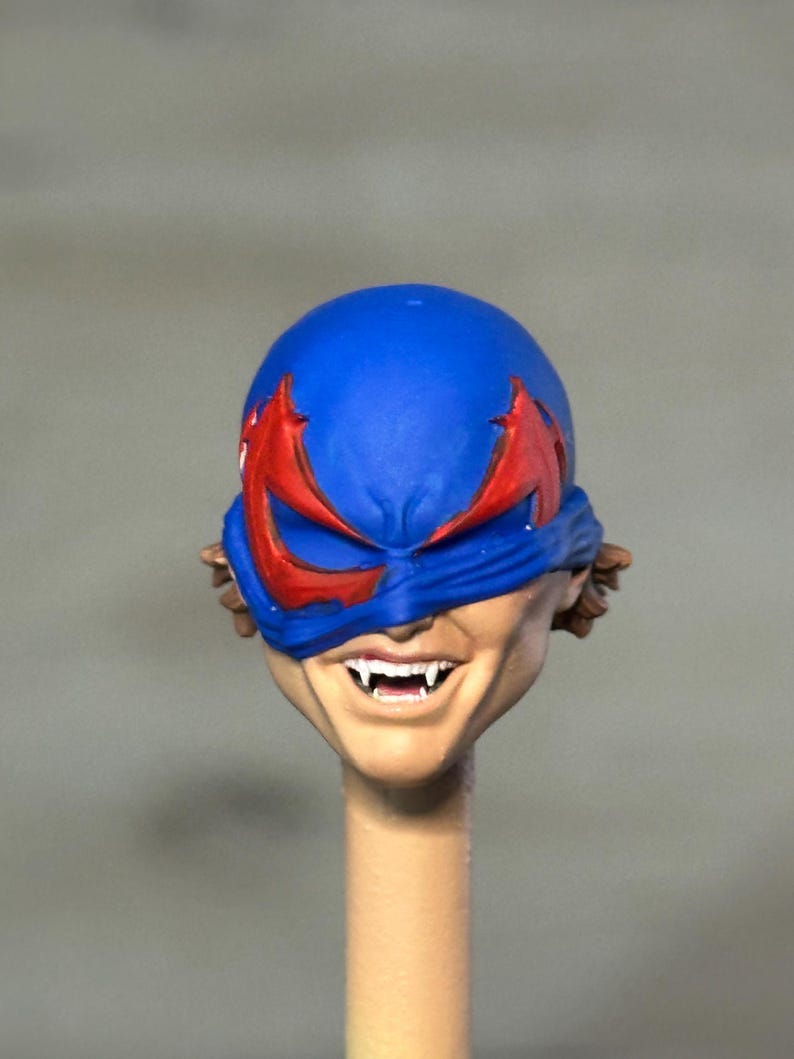 May include: A sculpted action figure head featuring a bright blue mask with red eye accents. The face has a light skin tone, a wide grin, and visible white fangs protruding from the mouth. Brown hair is partially visible on the sides of the head.