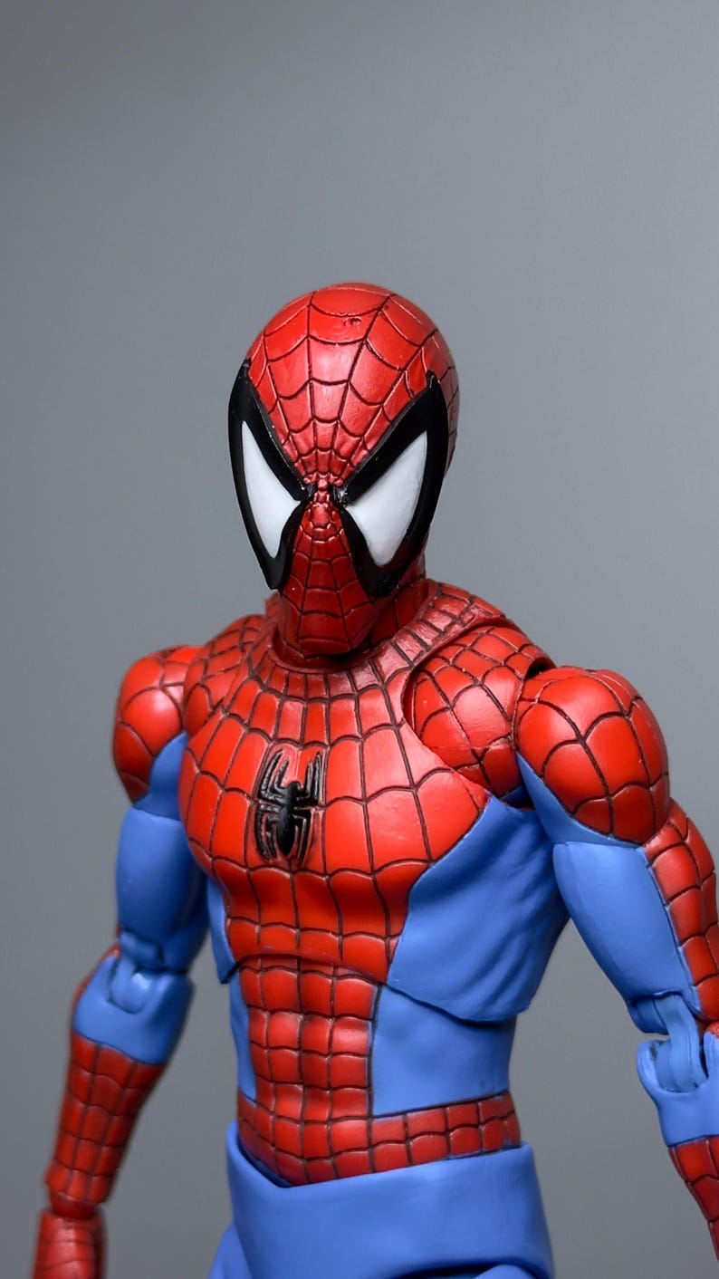 Puede incluir: Figura de acci&oacute;n detallada de Spider-Man, con su traje rojo y azul con un dise&ntilde;o de telara&ntilde;a. La figura tiene un emblema de ara&ntilde;a negra en el pecho y lentes oculares blancas. Fondo gris.