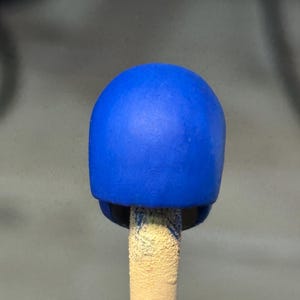 May include: A small, smooth, solid blue helmet-shaped object mounted on a textured, light-colored cylindrical stick. The blue surface has a matte finish and a rounded, dome-like top, appearing as a miniature component or a scale model accessory.