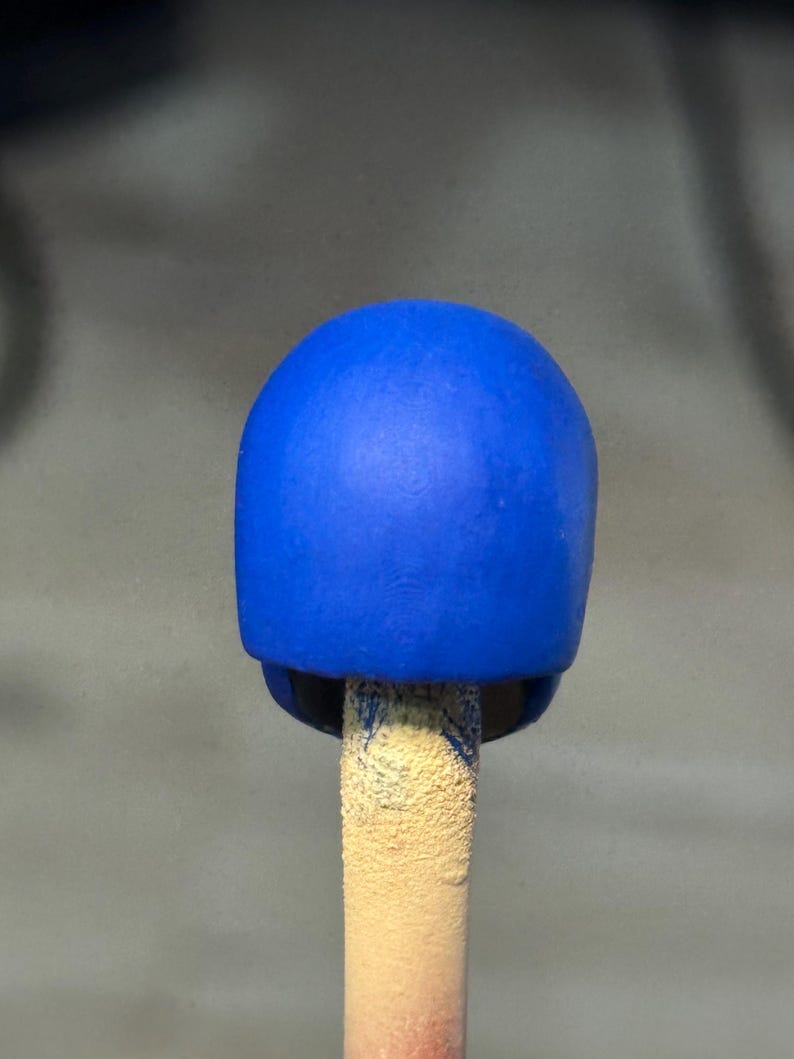 May include: A small, smooth, solid blue helmet-shaped object mounted on a textured, light-colored cylindrical stick. The blue surface has a matte finish and a rounded, dome-like top, appearing as a miniature component or a scale model accessory.