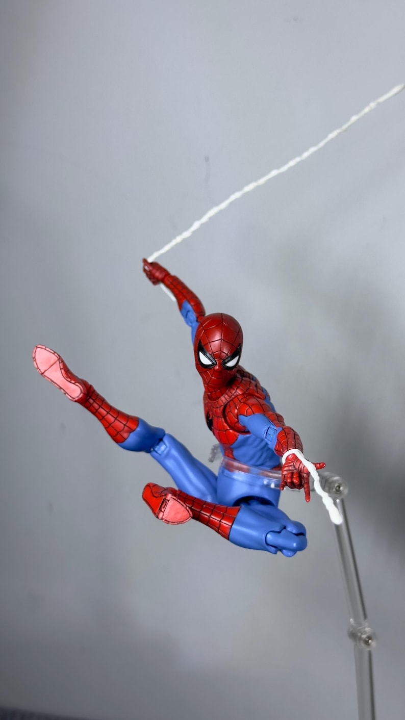 Puede incluir: Figura de acci&oacute;n de Spider-Man, rojo y azul, en una pose din&aacute;mica, con una telara&ntilde;a blanca que se extiende desde la mano de la figura. La figura est&aacute; en el aire, con una pierna extendida. La figura est&aacute; sobre un soporte transparente.