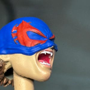 May include: A sculpted action figure head featuring a blue mask with red eye accents. The face has a light skin tone, an open mouth revealing white teeth and a red interior, and visible brown hair at the base of the neck, presented on a cylindrical stand.