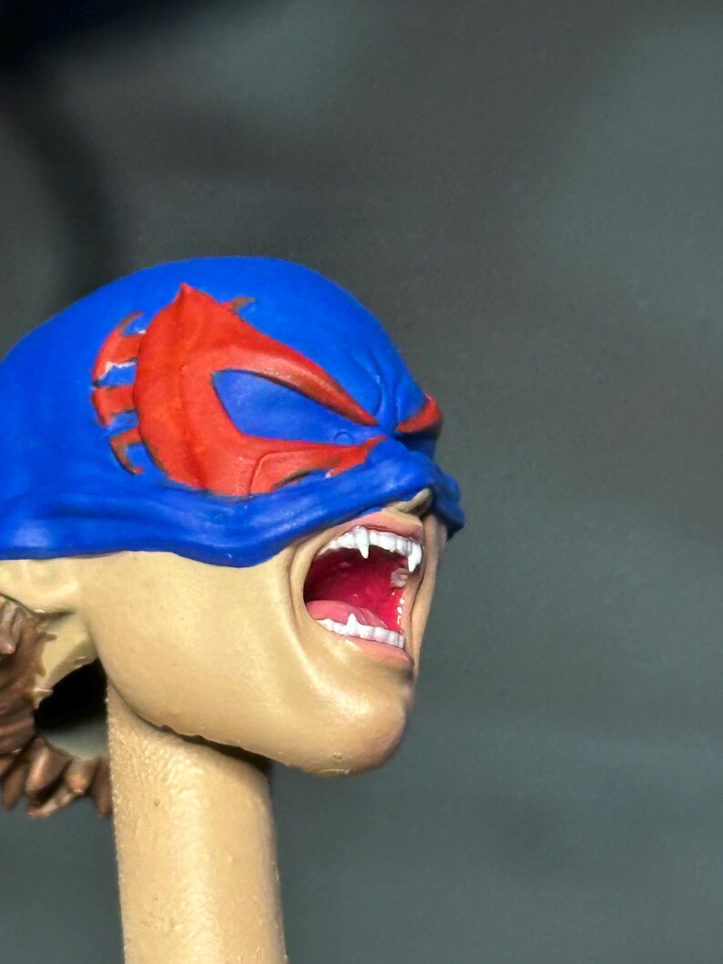 May include: A sculpted action figure head featuring a blue mask with red eye accents. The face has a light skin tone, an open mouth revealing white teeth and a red interior, and visible brown hair at the base of the neck, presented on a cylindrical stand.