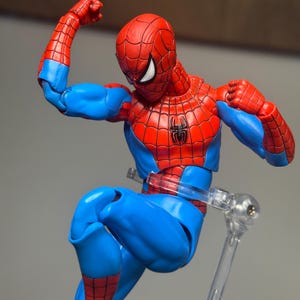 May include: A Spider-Man action figure in a dynamic pose, featuring a red and blue costume with a spider emblem on the chest. The figure is supported by a clear stand, showcasing the character's iconic design and colors.