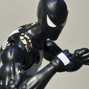 May include: A black action figure with a smooth, matte finish, featuring white eye lenses and a white chest emblem. The figure is posed in a fighting stance with clenched fists, showing visible paint wear and scuffing on the shoulder and arm joints.