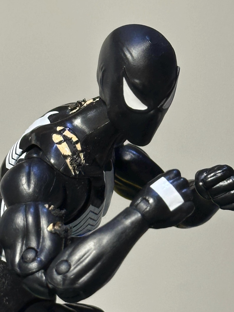 May include: A black action figure with a smooth, matte finish, featuring white eye lenses and a white chest emblem. The figure is posed in a fighting stance with clenched fists, showing visible paint wear and scuffing on the shoulder and arm joints.