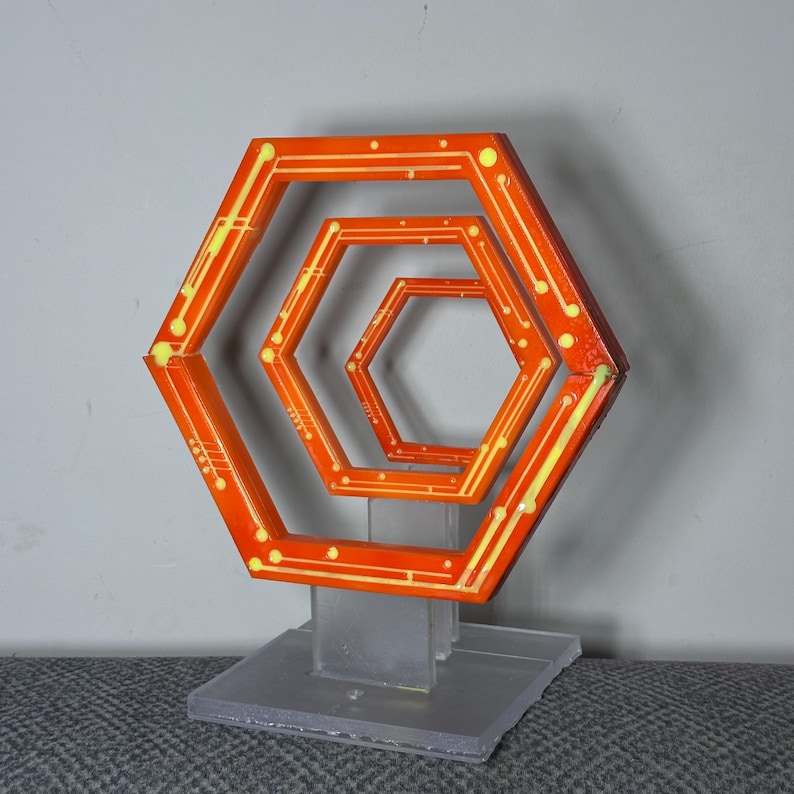 May include: An orange and yellow geometric sculpture featuring nested hexagonal shapes. The sculpture has a clear base and a series of orange hexagons with yellow accents and white dots. The design creates a three-dimensional effect.