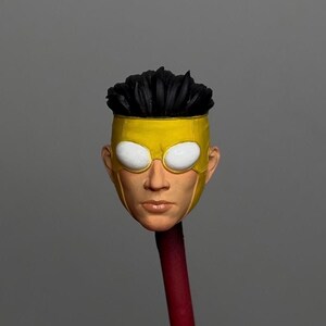 Può includere: Una scultura dettagliata della testa di un personaggio di supereroe. La figura presenta una maschera gialla con forme di occhi bianchi, capelli neri e un tono della pelle realistico. La testa è montata su un bastoncino rosso e marrone.