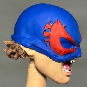 May include: A sculpted figurine head featuring a bright blue mask with a prominent red emblem over the eyes. The figure has light skin, visible white teeth in an open mouth, and brown hair styled in a short, textured cut at the back of the neck.