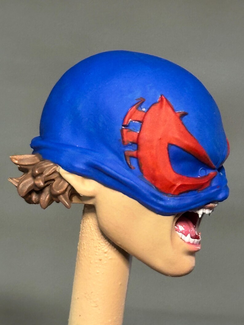 May include: A sculpted figurine head featuring a bright blue mask with a prominent red emblem over the eyes. The figure has light skin, visible white teeth in an open mouth, and brown hair styled in a short, textured cut at the back of the neck.