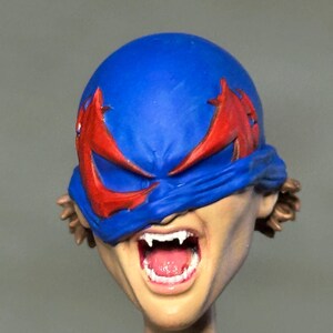 May include: A sculpted action figure head featuring a blue mask with red eye markings covering the upper face. The mouth is open in a wide expression, revealing white fangs and a pink tongue. Brown hair is visible at the sides of the head.