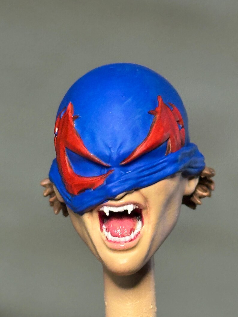 May include: A sculpted action figure head featuring a blue mask with red eye markings covering the upper face. The mouth is open in a wide expression, revealing white fangs and a pink tongue. Brown hair is visible at the sides of the head.
