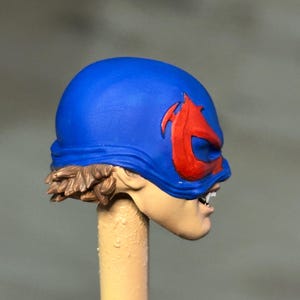 May include: A miniature figurine head featuring a bright blue mask with a prominent red emblem over the eyes. The figure has light skin tone, visible white teeth, and brown hair sculpted at the base of the mask, mounted on a beige cylindrical post.