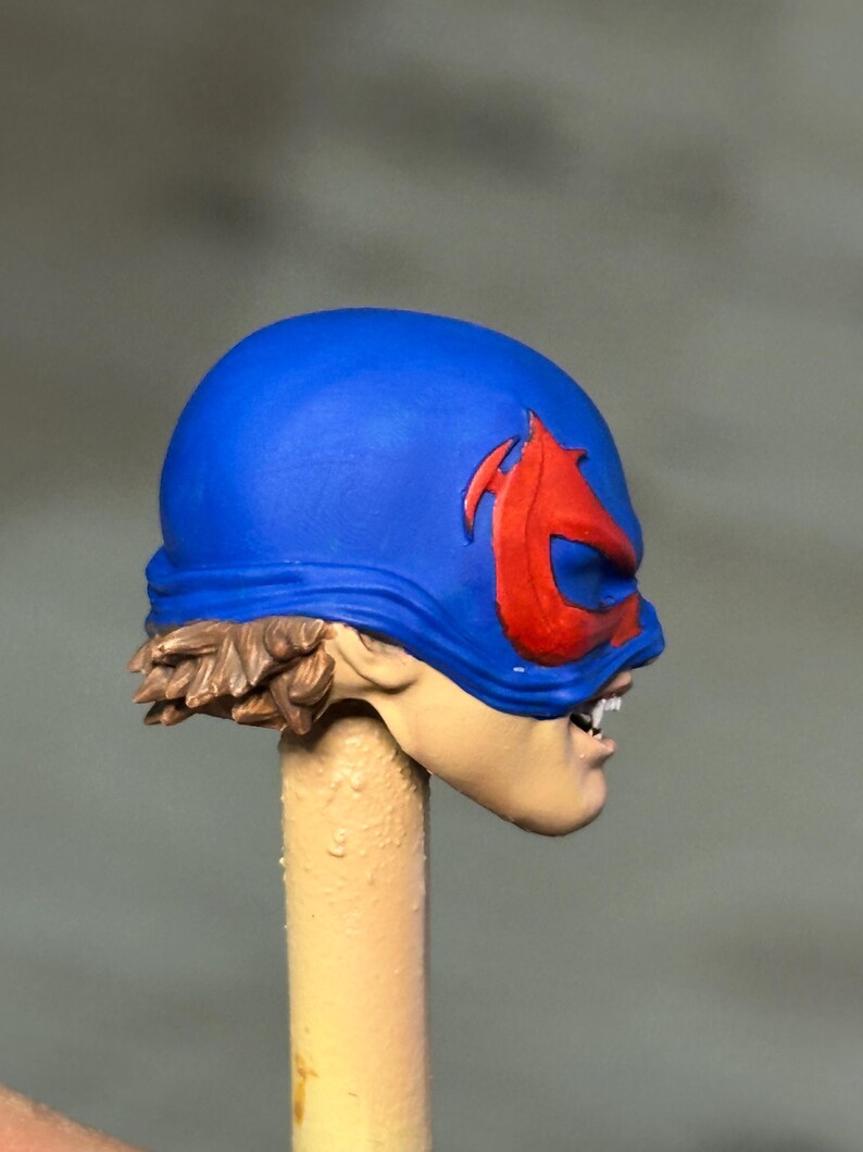 May include: A miniature figurine head featuring a bright blue mask with a prominent red emblem over the eyes. The figure has light skin tone, visible white teeth, and brown hair sculpted at the base of the mask, mounted on a beige cylindrical post.