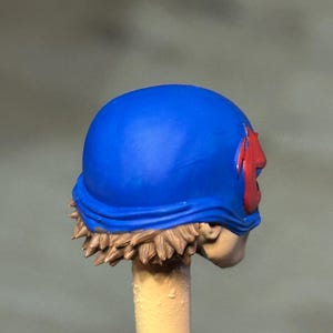 May include: A miniature figurine head featuring a bright blue helmet with a red emblem on the side. The figure has sculpted light brown hair visible beneath the helmet and a pale skin tone, mounted on a thin, light-colored cylindrical support rod.