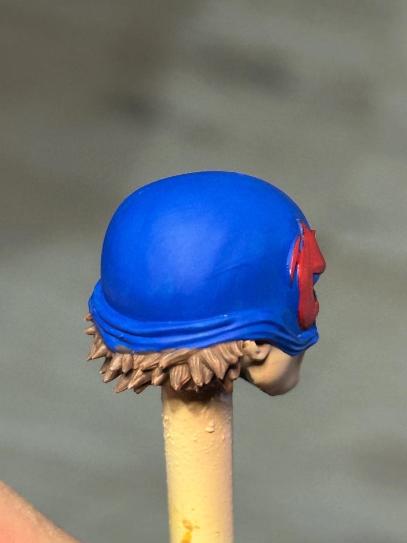 May include: A miniature figurine head featuring a bright blue helmet with a red emblem on the side. The figure has sculpted light brown hair visible beneath the helmet and a pale skin tone, mounted on a thin, light-colored cylindrical support rod.