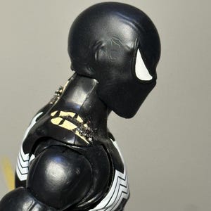 May include: A black action figure with a smooth, rounded head and a single white eye visible in profile. The figure features a black torso with white markings on the chest and shoulders, showing signs of wear with chipped paint revealing beige.