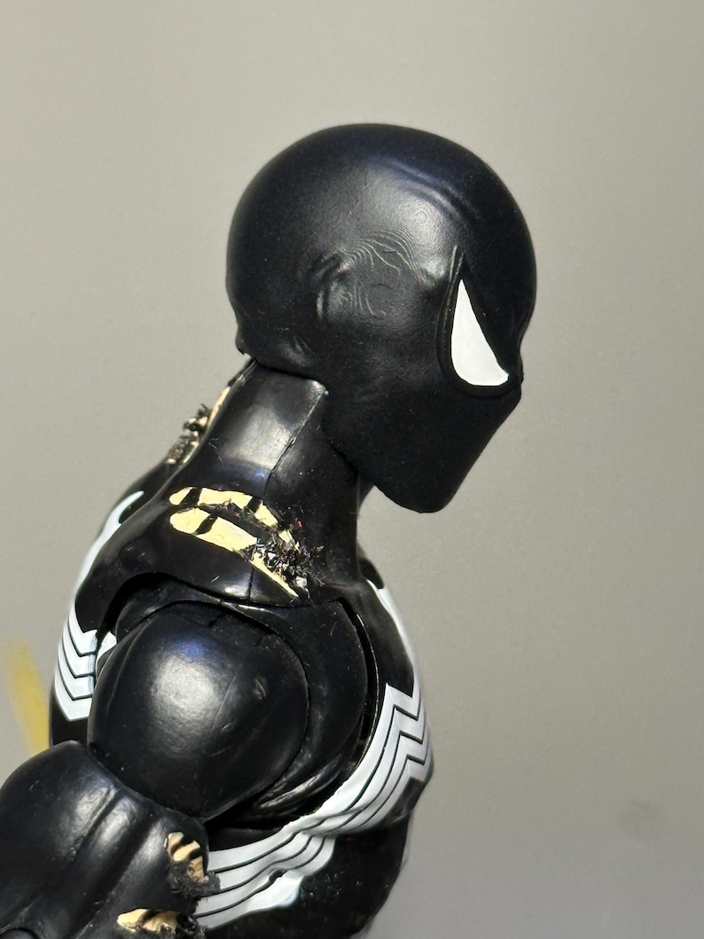 May include: A black action figure with a smooth, rounded head and a single white eye visible in profile. The figure features a black torso with white markings on the chest and shoulders, showing signs of wear with chipped paint revealing beige.