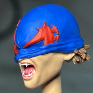 May include: A painted miniature head sculpt featuring a blue mask with red accents covering the eyes. The face has a light skin tone, an open mouth with visible white teeth and a pink tongue, and brown hair visible at the back of the head.