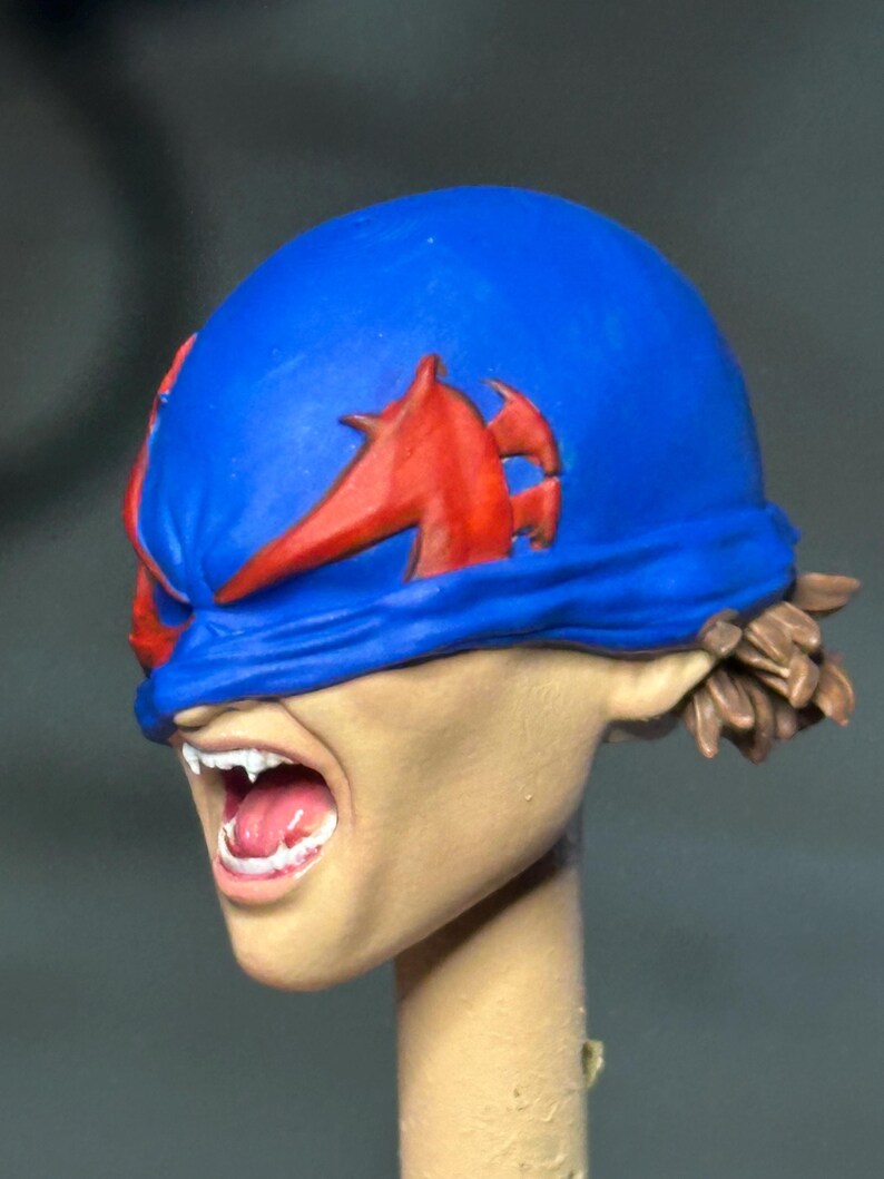 May include: A painted miniature head sculpt featuring a blue mask with red accents covering the eyes. The face has a light skin tone, an open mouth with visible white teeth and a pink tongue, and brown hair visible at the back of the head.